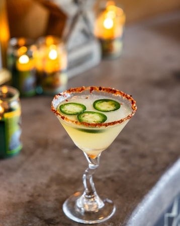 Chili-salt-rimmed jalapeño cocktail in a twisted-stem martini glass, topped with three jalapeño slices and set on a bar countertop with warm candles glowing in the background.