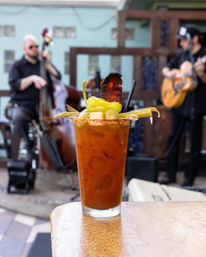 Spicy Bloody Mary in a tall glass with chili-rimmed edge, pickled pepper, green olives, celery and bacon garnish on a wooden patio table, blurred live acoustic duo playing upright bass and guitar in the outdoor background