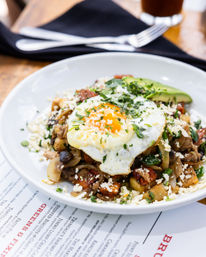 Close-up of a restaurant brunch plate: crispy potato hash with sautéed onions, mushrooms and herbs, topped with a sunny-side-up egg, crumbled cheese and avocado slices.