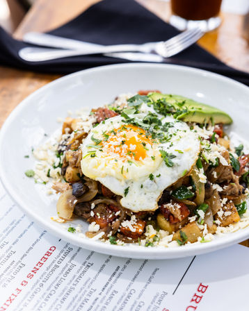 Close-up of a restaurant brunch plate: crispy potato hash with sautéed onions, mushrooms and herbs, topped with a sunny-side-up egg, crumbled cheese and avocado slices.