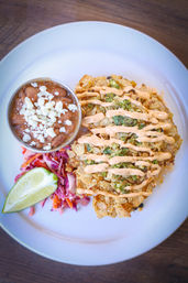 Top-down photo of a Mexican-style plate: chip-crusted crispy fritter drizzled with spicy crema and green salsa, served with refried beans topped with crumbled cheese, purple cabbage slaw and a lime wedge on a white plate.