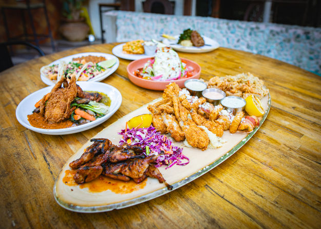 Crispy seafood and grill platter with fried shrimp and oysters, glazed grilled chicken, purple cabbage slaw, lemon wedges and three dipping sauces on an oval plate on a wooden restaurant patio table with extra dishes in the background.