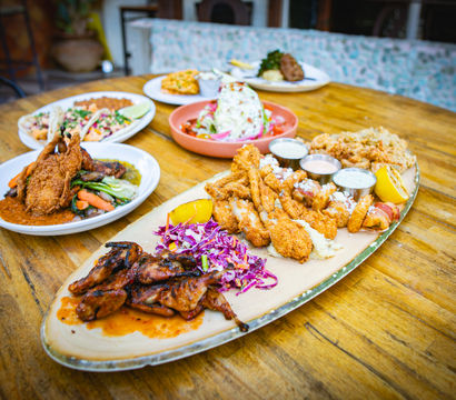Crispy seafood and grill platter with fried shrimp and oysters, glazed grilled chicken, purple cabbage slaw, lemon wedges and three dipping sauces on an oval plate on a wooden restaurant patio table with extra dishes in the background.