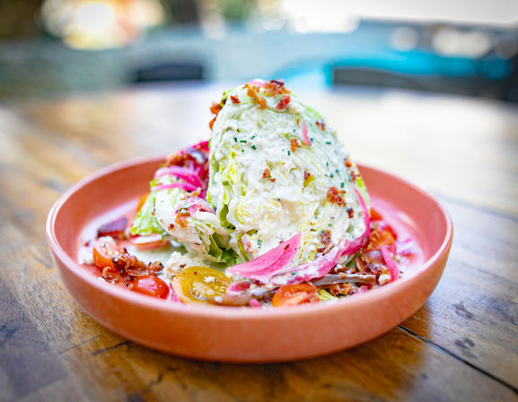 Creamy iceberg wedge salad topped with bacon bits, pickled red onions, cherry tomatoes and chives, served on a terracotta plate on an outdoor wooden table