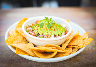 Vibrant bowl of pico de gallo topped with fanned avocado, cilantro and chili flakes, served with crispy tortilla chips on a wooden table — fresh Mexican-style appetizer.