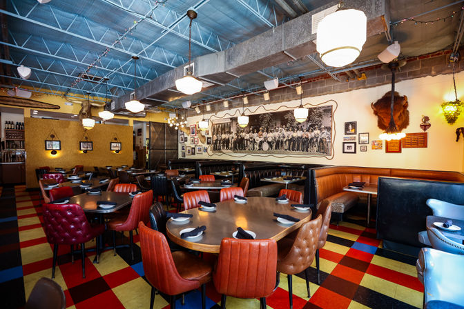 Vibrant retro-style restaurant interior with multicolor checkered floor, tufted leather chairs and curved booths around set wooden tables, hanging pendant lights and vintage photos including a large black-and-white mural on the wall.