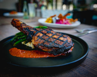 Grilled bone-in pork chop with charred crosshatch grill marks on a black plate, red pepper sauce and green beans on a restaurant table