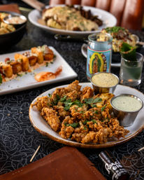 Gastropub-style appetizer: plate of crispy golden fried chicken bites garnished with herbs and grilled lemon, served with two creamy dipping sauces on a patterned restaurant table with small plates, a votive candle, and a cocktail nearby.