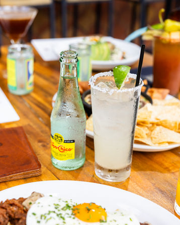 Salt-rimmed margarita with a lime wedge and straw beside a chilled sparkling water bottle on a wooden table, surrounded by tortilla chips and a sunny-side-up egg plate in a bright casual brunch scene.