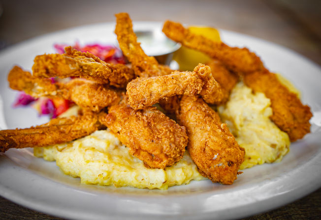 Crispy Southern-style fried chicken strips stacked on creamy cheesy grits with a lemon wedge, dipping sauce and colorful coleslaw on a white plate