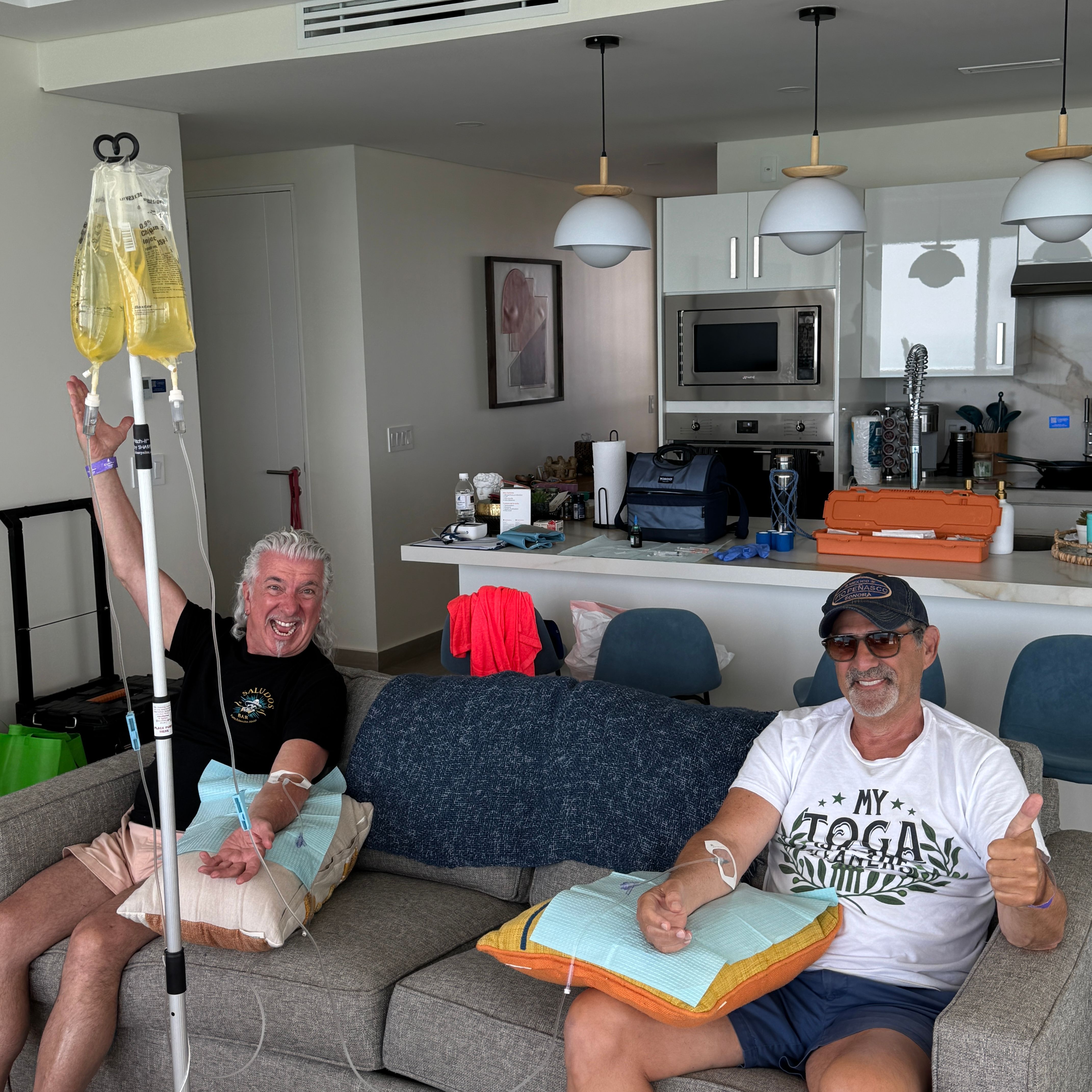 Two smiling men lounging on a gray couch in a bright modern apartment living room with open kitchen, each receiving yellow IV vitamin drips; one holds the IV pole and the other gives a thumbs-up, cushions on their laps for comfort.