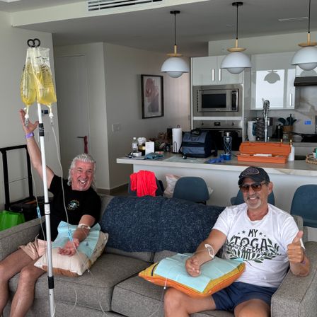 Two smiling men lounging on a gray couch in a bright modern apartment living room with open kitchen, each receiving yellow IV vitamin drips; one holds the IV pole and the other gives a thumbs-up, cushions on their laps for comfort.