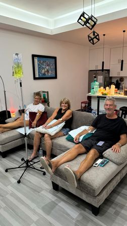 Three adults reclining on a gray sectional in a modern living room receiving IV drips from a pole, smiling and relaxed with a kitchen island visible in the background — at-home IV therapy scene.