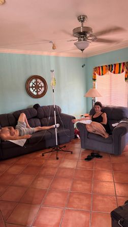 Bright home living room with terracotta tile floor and turquoise walls, two gray couches and ceiling fan; a person reclining and another seated connected to an IV pole in the center, colorful window valance and lamp nearby.