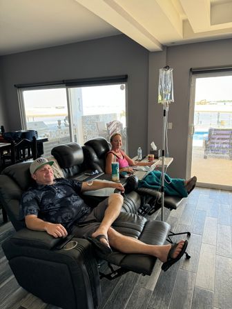 Two adults relaxing in black leather recliners in a bright coastal living room, one receiving IV hydration from a hanging bag while the other uses a laptop; sliding glass doors open to a sunny patio and water view.