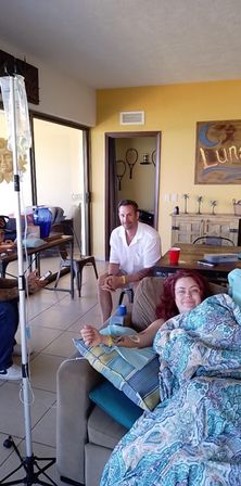 Sunlit living room with sliding glass door: a person reclines on a sofa wrapped in a teal paisley blanket connected to an IV drip, while a friend sits nearby by a yellow accent wall and dining table.