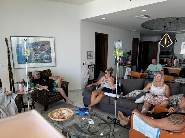 Relaxed group of adults in a sunlit coastal-style living room of a vacation condo, seated on sofas and chairs receiving IV hydration drips from upright poles.