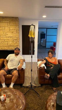 Two smiling people seated in leather armchairs in a cozy living-room wellness space receiving yellow IV drip bags from a central pole, with a stone accent wall and ottoman visible.