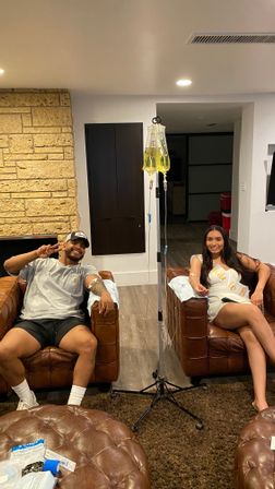 Two adults relaxed in brown leather armchairs in a cozy living room, smiling while receiving yellow IV drips from a central IV pole — ottoman and medical supplies visible, suggesting an at-home IV therapy wellness session.