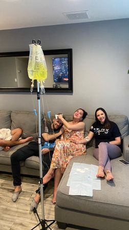 Three adults relaxing on a gray living-room sectional receiving at-home IV hydration — yellow IV bags on a pole, medical supplies on the ottoman, playful smiling moment.