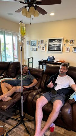 Two adults reclining on a brown leather sofa in a residential living room receiving IV drips from a standing pole with a yellow bag; one gives a thumbs-up, framed photos and a sliding glass door visible.