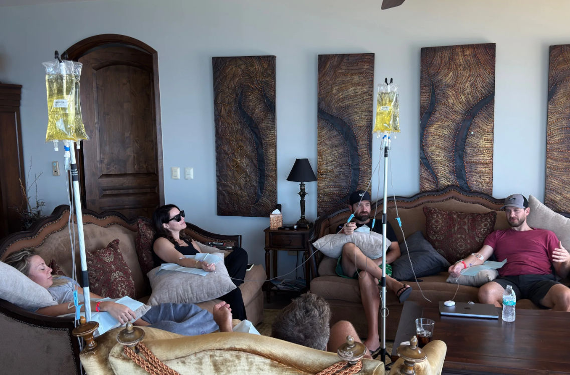 Group IV drip therapy session in a plush living room — several adults reclining on sofas with IV bags on poles, decorative wooden wall panels, a side table lamp and coffee table visible.