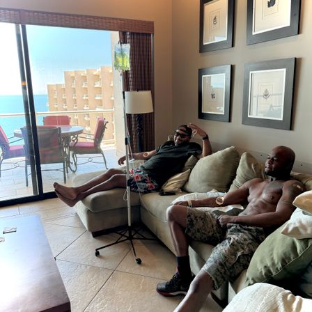 Two men relaxing on a beige sectional in an ocean-view condo — one in sunglasses and floral shorts flashing a peace sign, the other shirtless with an IV drip, sliding glass door open to a balcony with red patio chairs and blue sea