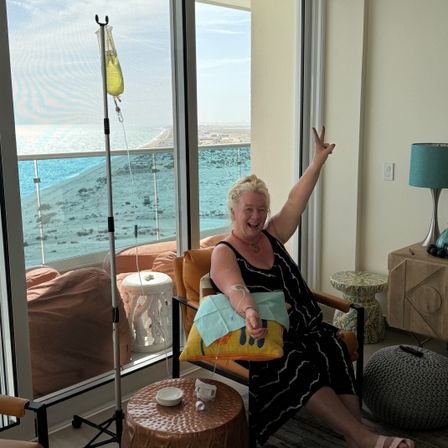 Cheerful woman flashing a peace sign while receiving an IV drip in a bright oceanfront condo overlooking a sandy beach and balcony.