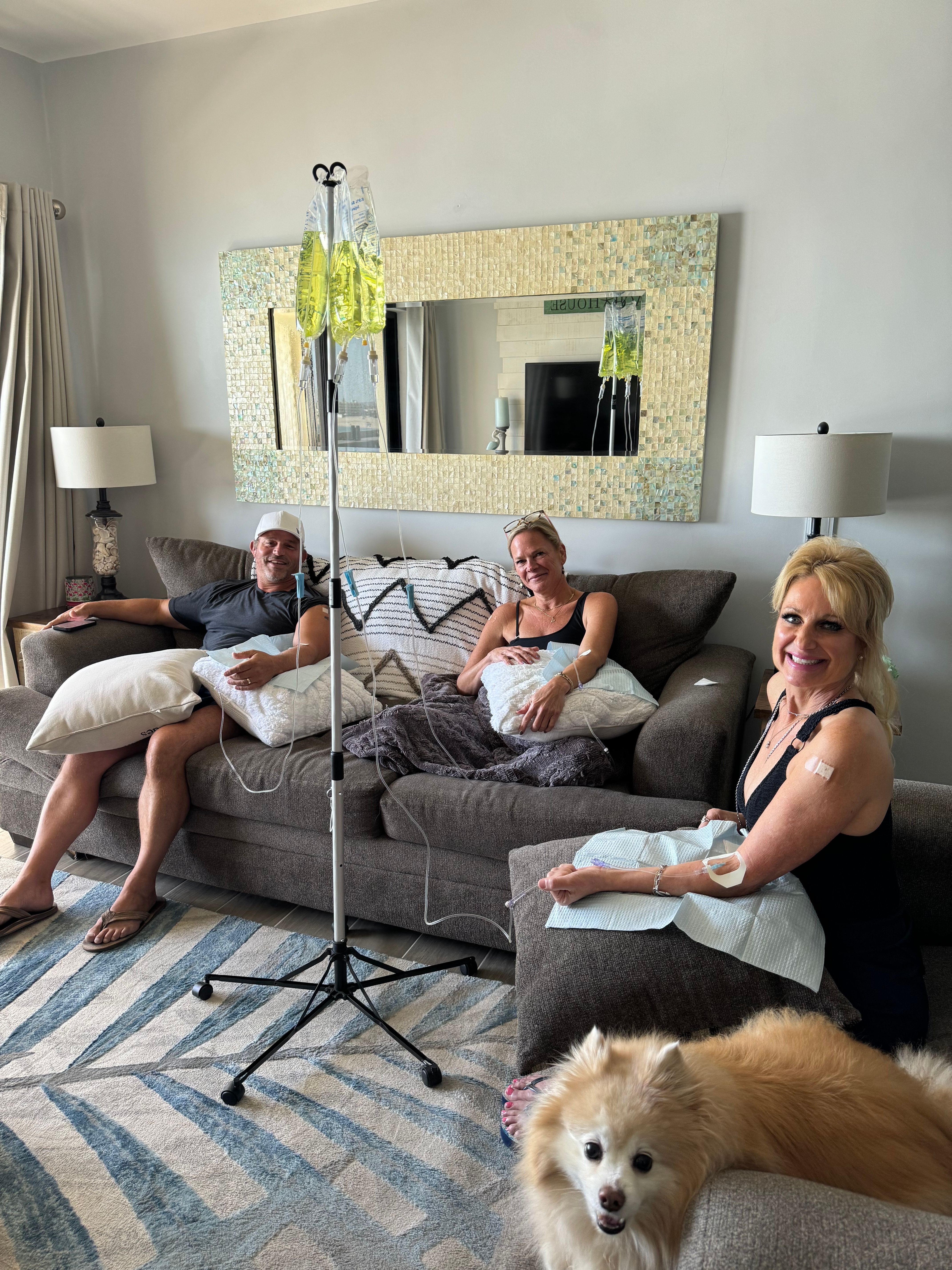 Three adults relaxing on a living-room couch receiving IV infusions from a nearby IV stand, smiling woman with a bandage on her arm in the foreground and a fluffy dog on the ottoman; mosaic mirror and lamps in the background — at-home IV therapy session.