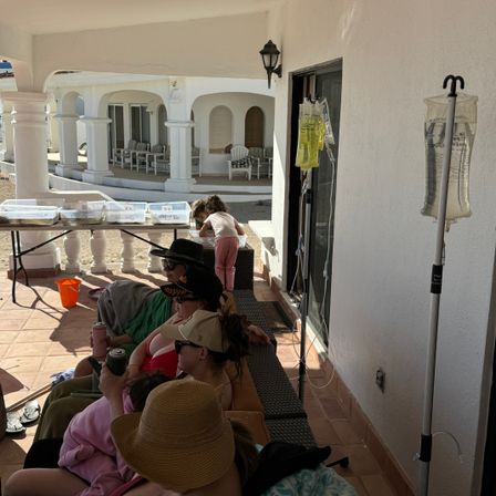 Group relaxing on a sunny white-stucco patio with arched columns and wicker seating, children playing at a table, and two IV poles with hanging infusion bags by the sliding door
