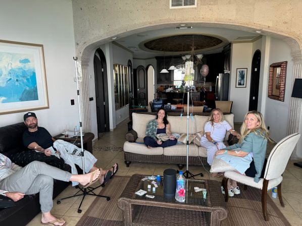 Group of four adults lounging in a bright vacation rental living room with wicker sofa and chairs, tiled floors, arched doorway to an open kitchen, coffee table and IV infusion stands in the center.