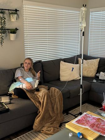 Person on a gray sectional in a cozy living room receiving an IV infusion, thumbs up, wrapped in a tan blanket with decorative pillows, IV pole and saline bag beside the sofa, potted plants and closed blinds in background.