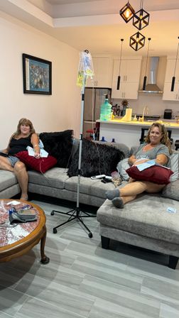 Two women relaxing on a gray sectional sofa receiving IV therapy with an IV drip stand between them, modern open-plan kitchen and pendant lights in the background.