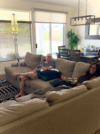 Two smiling adults reclining on a sunlit living room sofa with an IV bag on a stand and tubing attached, thumbs-up vibe, modern dining area and potted plant in the background.