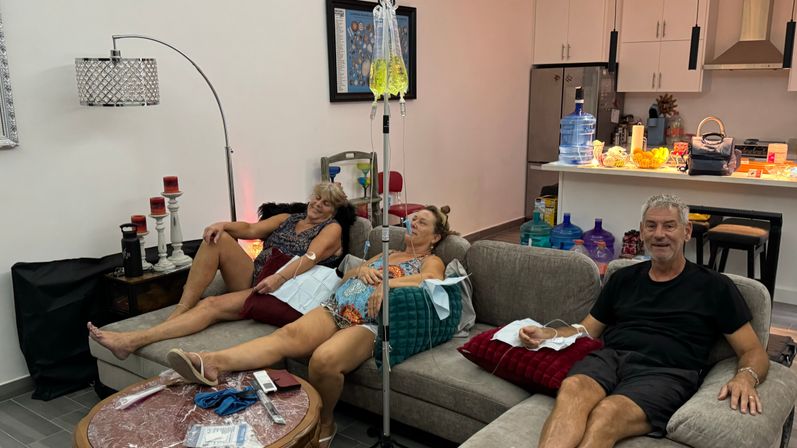 Three adults lounging on a living-room sofa in a modern apartment receiving IV vitamin drips from a nearby stand — relaxed at-home wellness session.
