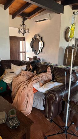 Two people wrapped in pink blankets sitting on a brown leather sofa in a rustic, tile-floored living room giving thumbs up; an IV pole with a yellow IV bag and tubing stands nearby under a wood-beamed ceiling with a decorative mirror and chandelier.