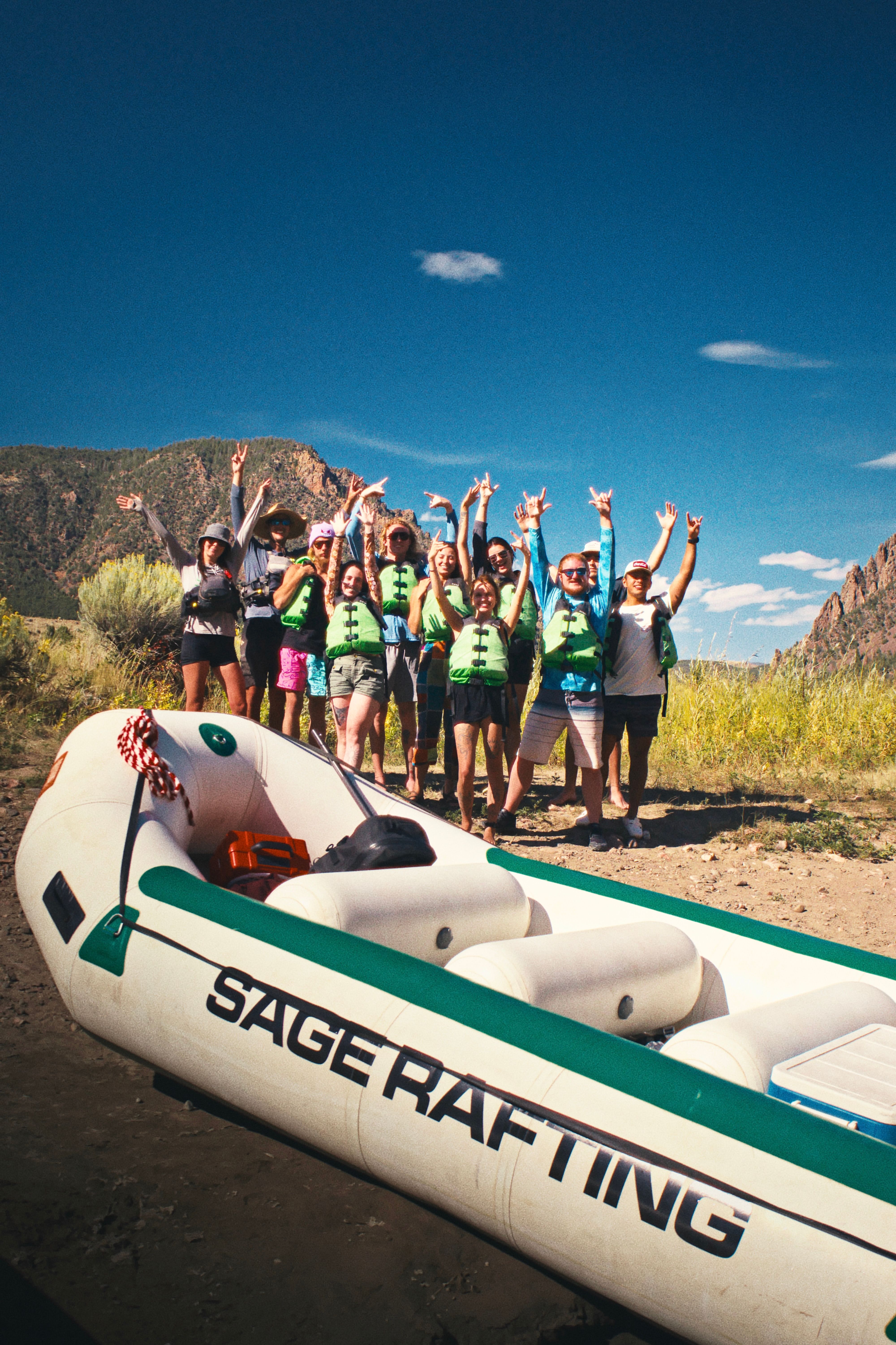 Cheerful group of rafters wearing green life jackets cheering on a rocky riverbank beside a white-and-green inflatable raft, with canyon cliffs and a clear blue sky in the background