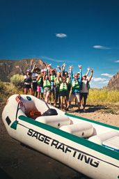 Cheerful group of rafters wearing green life jackets cheering on a rocky riverbank beside a white-and-green inflatable raft, with canyon cliffs and a clear blue sky in the background