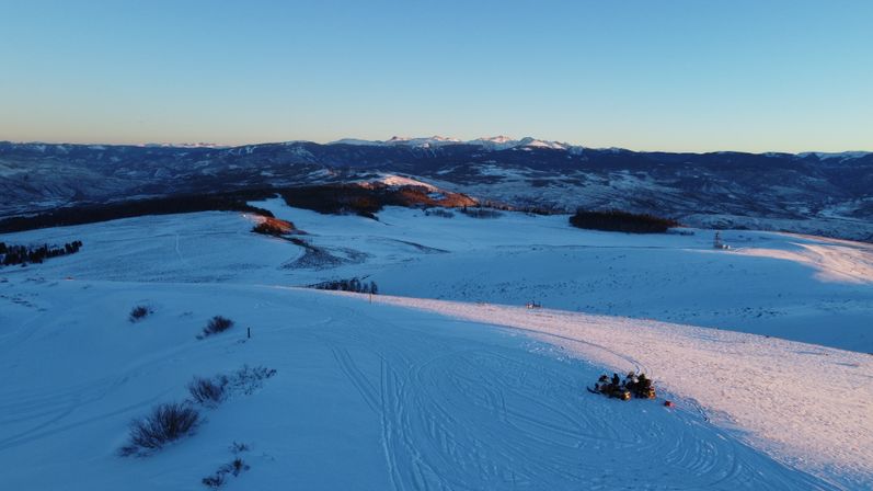 Aerial winter landscape of snow-covered alpine ridge at sunrise with distant mountain peaks and a small group of snowmobilers on a sunlit slope.
