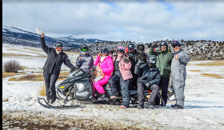 Eight people in helmets and colorful snow suits posing on and beside a long snowmobile in a snowy mountain meadow, a fun winter snowmobile adventure in a scenic mountain valley.