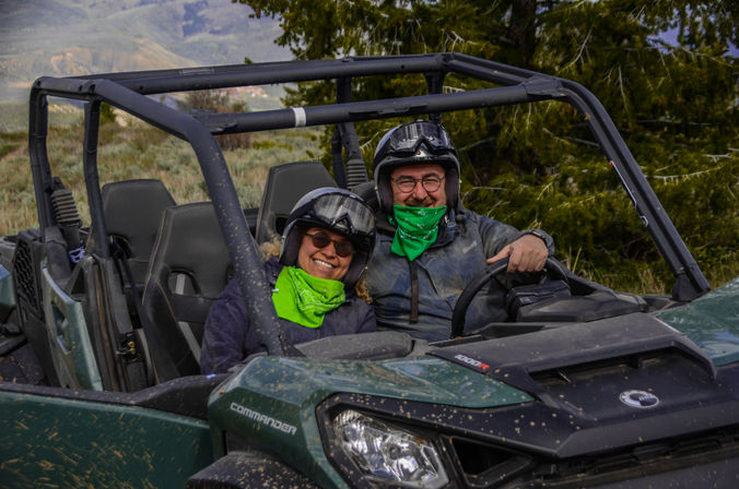 Two riders wearing helmets and bright green bandanas smile in a muddy green UTV on an off-road mountain trail with pine trees and distant hills.