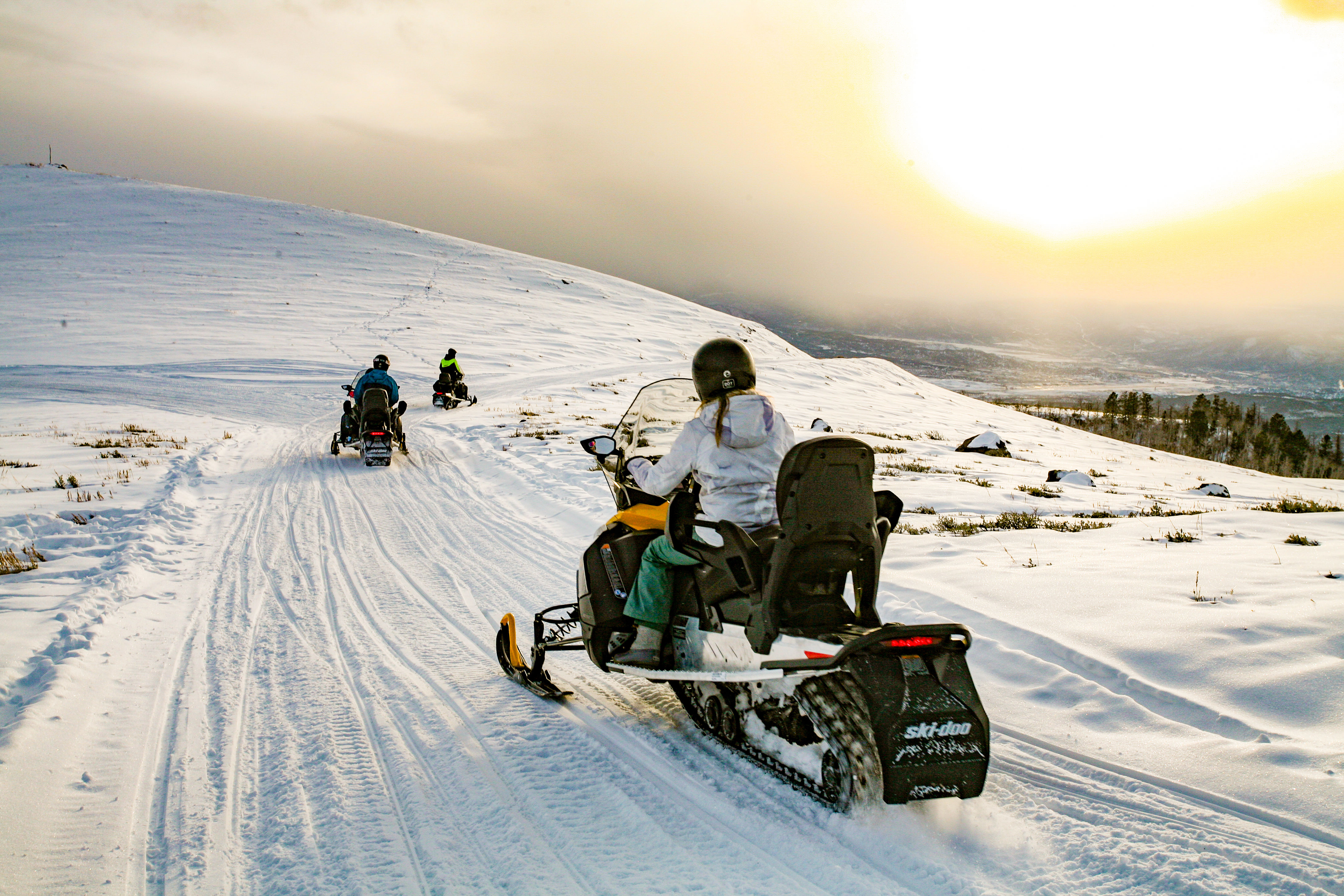 Three snowmobile riders on a groomed snowy trail across a sunlit mountain hillside at sunset, scenic winter landscape with a distant valley.