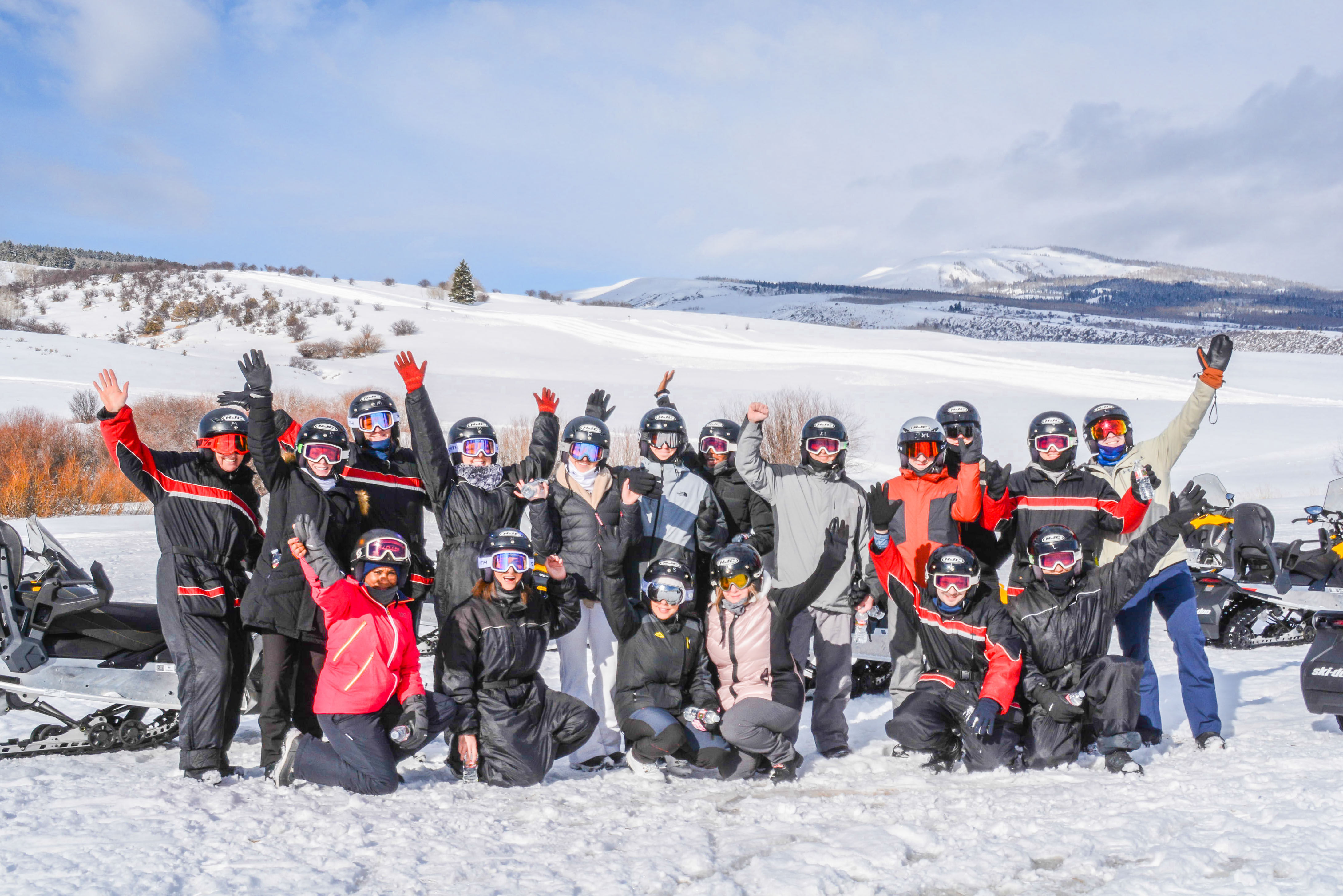 Cheerful group in helmets, goggles and winter suits posing with snowmobiles on a sunny snowy mountain landscape — winter snowmobile tour.