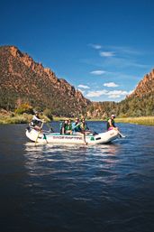 Group river rafting trip through a sunlit rocky canyon with pine-covered cliffs, clear blue sky, and calm water