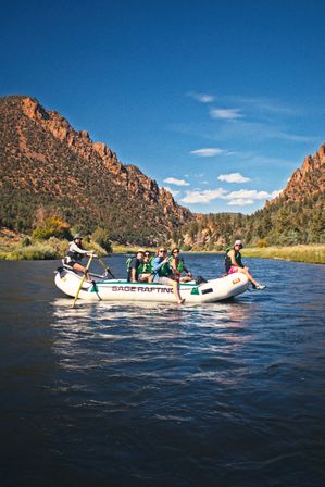 Group river rafting trip through a sunlit rocky canyon with pine-covered cliffs, clear blue sky, and calm water