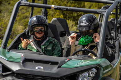 Two helmeted riders in a green off-road UTV on a brushy hillside; passenger flashes a shaka sign while the driver wears a green bandana.
