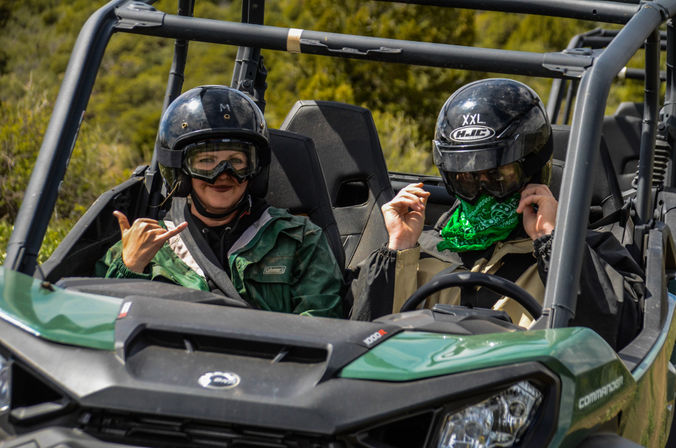 Two helmeted riders in a green off-road UTV on a brushy hillside; passenger flashes a shaka sign while the driver wears a green bandana.