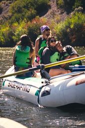 Four people in green life jackets relaxing on a white inflatable raft with oars, drifting on a sunlit tree-lined river — summer river rafting adventure.