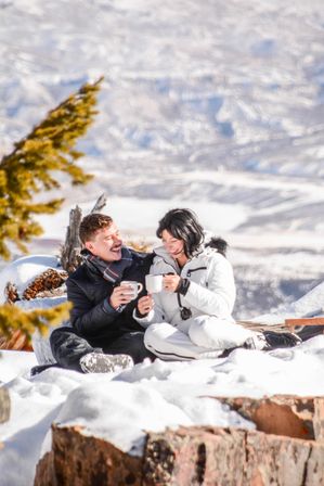 Smiling couple in winter jackets sitting on snow-covered logs, clinking mugs of hot drink amid snowy alpine mountain scenery.