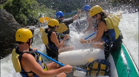 Group whitewater rafting through rocky river rapids, wearing yellow helmets and life vests as blue paddles cut through splashing water.